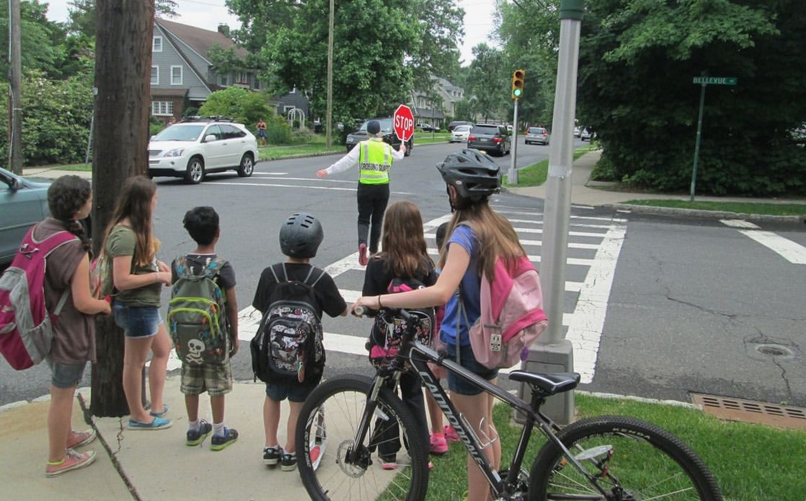 Group of kids walking and on bicycles waiting for the crossing guard at a cross walk.